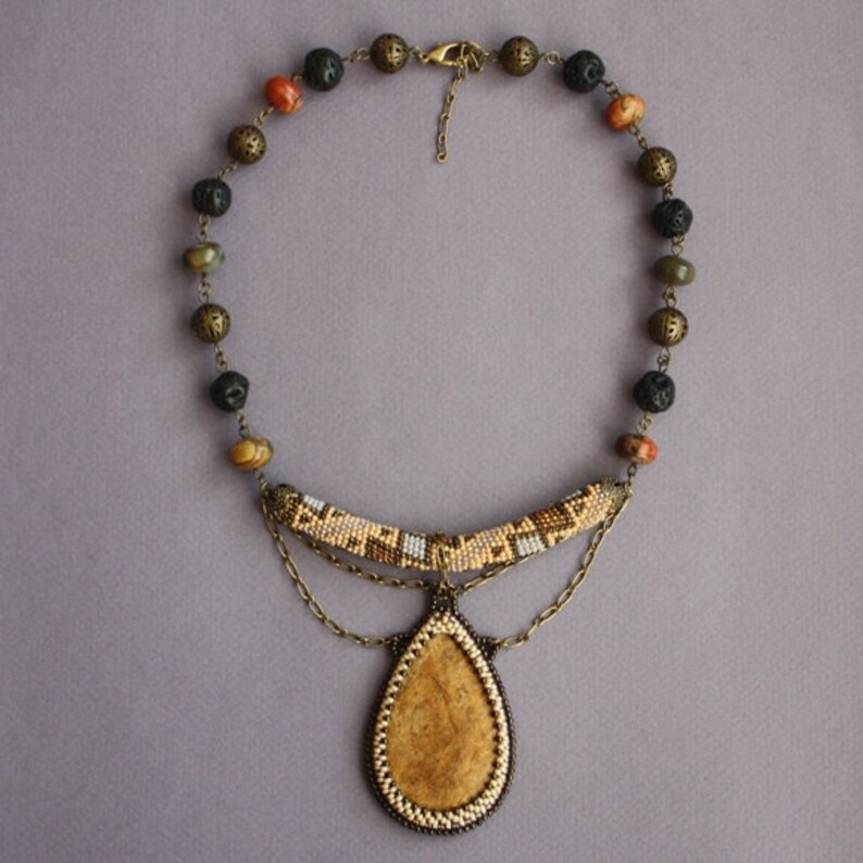 May include: A beaded necklace with a teardrop-shaped pendant. The pendant is made of a light brown material and is surrounded by a beaded border. The necklace is made of brown, black, and green beads.