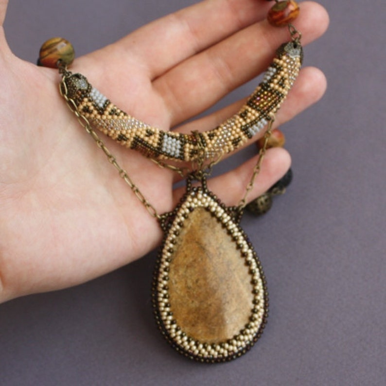 May include: A beaded necklace with a large teardrop-shaped stone pendant. The necklace is made with brown, beige, and white beads and has a chain with a small charm. The stone is a light brown color.