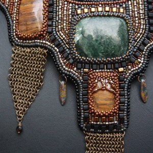May include: A beaded necklace featuring a large green stone at its centre, surrounded by smaller brown and black beads. The necklace has two gold chain tassels hanging from the bottom.