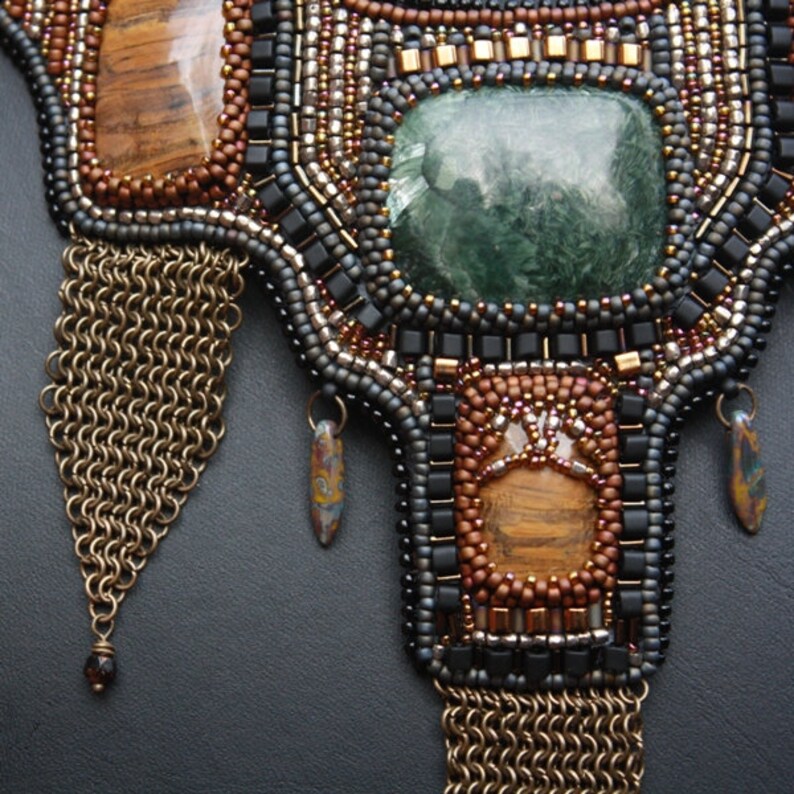 May include: A beaded necklace featuring a large green stone at its centre, surrounded by smaller brown and black beads. The necklace has two gold chain tassels hanging from the bottom.