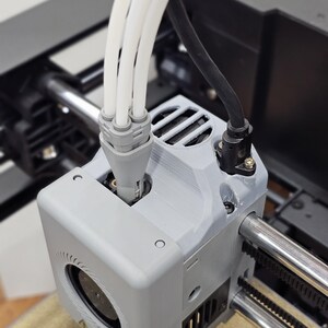 May include: A 3D printer print head assembly featuring a light gray housing with a circular fan vent. Three white filament tubes connect to a gray coupling on top, while a black cable connects to the side. The unit is mounted on metal rails.