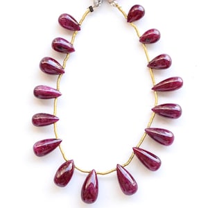 May include: A necklace featuring teardrop-shaped, deep red gemstones strung on a gold-colored wire. The gemstones are evenly spaced and have a polished, glossy appearance. The necklace is displayed against a white background.