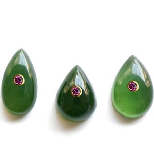 May include: Three teardrop-shaped green jade cabochons, each featuring a single small, round, faceted pink gemstone set in a gold-toned bezel at the center. The stones have a smooth, polished surface and a deep, translucent green color.