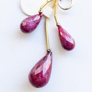 May include: A pendant with three faceted, teardrop-shaped, deep magenta gemstones. The gems are suspended from gold-coloured metal rods and a silver ring, with a red string detail. A white tag is attached.