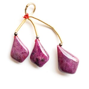 Puede incluir: Un colgante con tres gemas magenta pulidas en forma de lágrima. Las piedras están suspendidas de alambres dorados y un pequeño anillo dorado, conectados con hilo rojo. Las gemas tienen un acabado brillante.