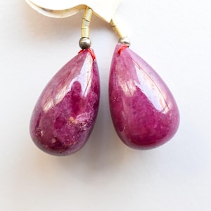 Könnte beinhalten: Ein Paar tropfenförmige Ohrringe in tiefem Magenta. Die polierten Edelsteine hängen an einem dünnen, goldfarbenen Draht mit einer silbernen Perle und roten Faden Details. Die Ohrringe sind vor einem weißen Hintergrund platziert.