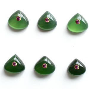 May include: Six smooth, triangular green cabochon stones arranged in two rows. Each stone features a small, round, vibrant pink gemstone set in a gold-colored bezel at the top center, creating a consistent decorative pattern across all pieces.