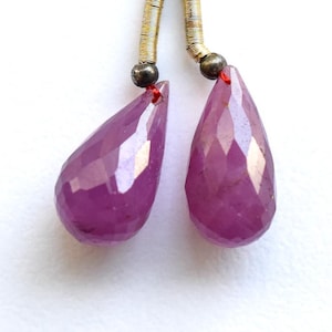 Puede incluir: Un par de gemas facetadas de color magenta en forma de lágrima suspendidas de un alambre dorado y un anillo de bronce. Las gemas están conectadas con un hilo rojo.