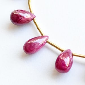 May include: A strand of teardrop-shaped, deep red ruby gemstones strung on a thin gold wire. The polished stones have a glossy finish and are evenly spaced, creating a necklace or jewelry component.