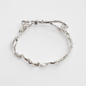 May include: A silver-colored bracelet with a twisted, organic design. The bracelet appears to be made of metal and has a flowing, irregular shape. The design features a knot-like element at the top and a continuous, wavy pattern throughout.