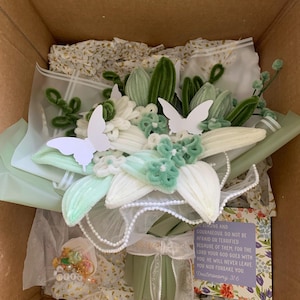 May include: A bouquet of light green and white chenille flowers wrapped in sage green paper with white lace trim. Two white paper butterflies are attached to the flowers. A small card with the text "Be strong and courageous. Do not be afraid or terrified because of them, for the Lord your God goes with you. He will never leave you nor forsake you. Deuteronomy 31:6" is included.