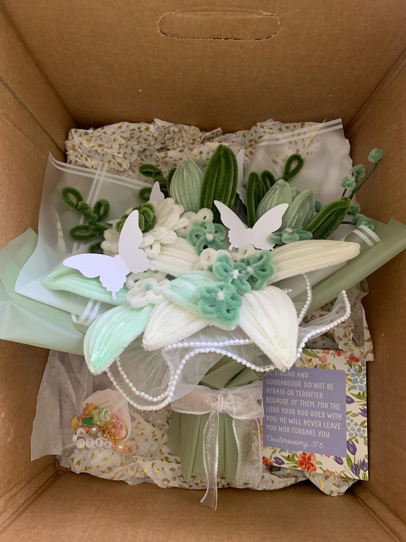 May include: A bouquet of light green and white chenille flowers wrapped in sage green paper with white lace trim. Two white paper butterflies are attached to the flowers. A small card with the text "Be strong and courageous. Do not be afraid or terrified because of them, for the Lord your God goes with you. He will never leave you nor forsake you. Deuteronomy 31:6" is included.