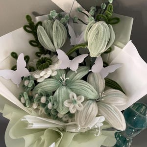 May include: A bouquet of handcrafted pipe cleaner flowers in shades of sage green and white, featuring various petal shapes and textures. The arrangement is accented with several white paper butterfly cutouts and wrapped in layers of light green paper.
