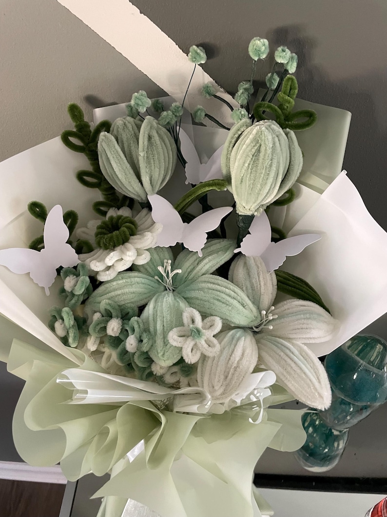 May include: A bouquet of handcrafted pipe cleaner flowers in shades of sage green and white, featuring various petal shapes and textures. The arrangement is accented with several white paper butterfly cutouts and wrapped in layers of light green paper.