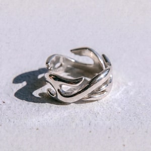 May include: A polished silver-toned ring featuring an open, fluid design with curved, flame-like metal segments that wrap around the band. The word "dusk." is printed in small, black, lowercase letters at the bottom of the image frame.