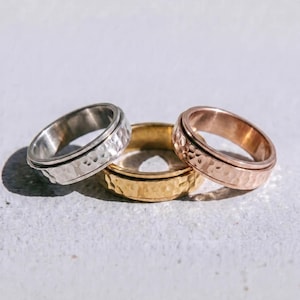 May include: Three hammered metal rings in silver, gold, and rose gold. The rings are displayed on a light-colored surface. The word "dusk." is printed in black at the bottom of the image. Jewelry.