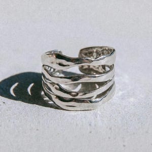 Könnte beinhalten: Ein silberfarbener Ring mit einem strukturierten, geschichteten Design. Der Ring hat ein breites Band mit offenem Rücken. Das Wort "dusk." ist unten im Bild schwarz gedruckt.