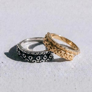 May include: Two rings, one silver and one gold, are displayed. The silver ring has a dark finish with evenly spaced circular indentations. The gold ring has a textured surface with similar indentations. The word "dusk." is printed at the bottom.