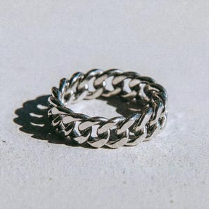 May include: A silver chain ring with a curb link design. The ring is made of a shiny, metallic material and is displayed on a light grey surface. The word "dusk." is printed in black at the bottom.
