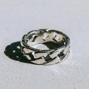 May include: A silver ring featuring a chain link pattern. The ring sits on a light grey surface. The word "dusk." is printed in black at the bottom of the image.