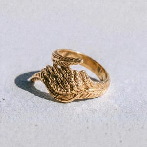 May include: A gold ring featuring a detailed feather design. The ring has a textured band and a swirling, open design. The word "dusk." is printed in a dark colour at the bottom of the image.