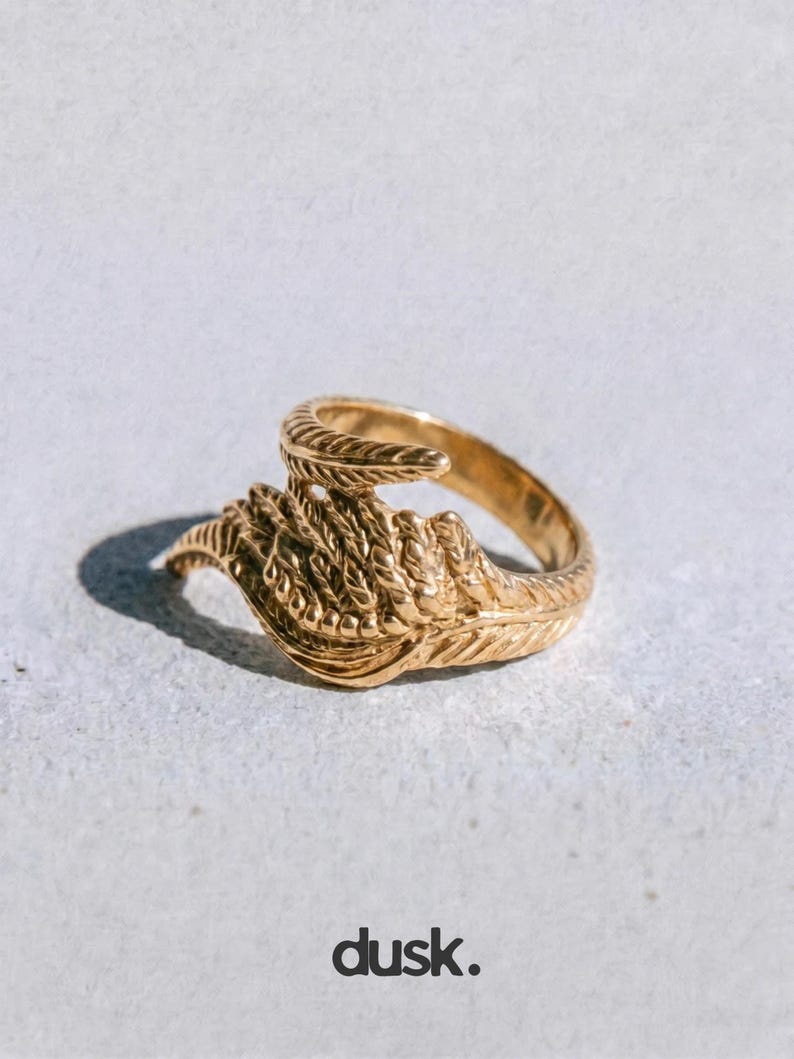 May include: A gold ring featuring a detailed feather design. The ring has a textured band and a swirling, open design. The word "dusk." is printed in a dark colour at the bottom of the image.