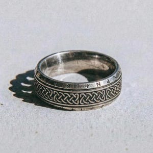 May include: A silver ring featuring a Celtic knot pattern and engraved symbols. The ring has a textured surface and is set against a light background. The word "dusk." is printed at the bottom of the image.