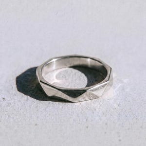 Puede incluir: Un anillo plateado con un diseño geométrico facetado. El anillo tiene una serie de superficies anguladas que captan la luz. La palabra "dusk." está impresa en negro en la parte inferior de la imagen.