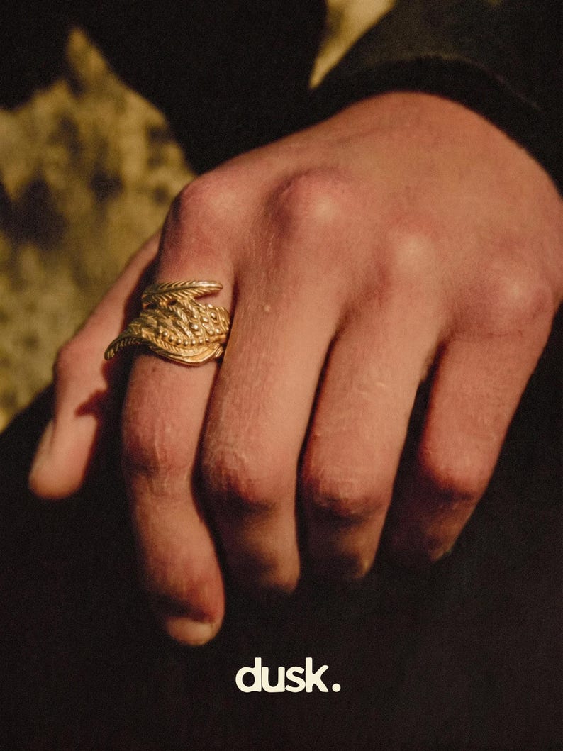 May include: A gold ring with a detailed, textured design is worn on a hand. The ring has a layered appearance. The word "dusk." is printed in white at the bottom of the image.