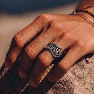 May include: A silver ring with a woven design is worn on a finger. The ring has a textured appearance and is set against a natural background. The word "dusk." is printed in white at the bottom of the image.