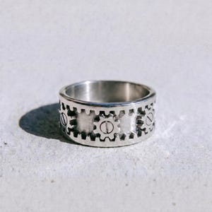 May include: A silver-toned metal ring featuring a band with a repeating gear-like pattern around the exterior. Each gear element includes a central horizontal line detail. The word dusk. is printed in black lowercase letters below the ring.