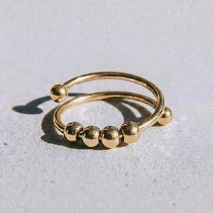 May include: A gold-colored ring with a spiral design and four small, round beads. The ring has a smooth, polished finish and is set against a light background. The word "dusk." is printed at the bottom.