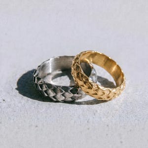May include: Two textured rings, one silver and one gold, are displayed on a light grey surface. The rings have a repeating diamond pattern. The word "dusk." is printed in a dark grey font at the bottom of the image.