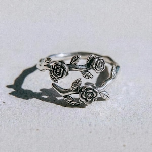 May include: A silver ring with a floral design, featuring three rose blossoms and leaves. The ring is shaped like a vine, with the roses and leaves wrapping around the band. The word "dusk." is printed at the bottom of the image.