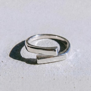 May include: A silver ring with a contemporary design. The ring has a band that encircles the finger with a rectangular bar detail. The word "dusk." is printed in black at the bottom of the image.