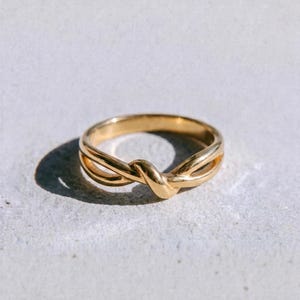 Op de afbeelding: Een gepolijste gouden ring met een centraal knoopontwerp waarbij twee banden elkaar kruisen en verstrengelen. De ring heeft een glad, reflecterend oppervlak en een afgerond profiel. Het woord dusk. staat in zwarte kleine letters onder de ring.