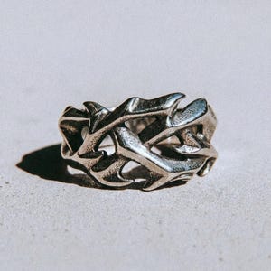 May include: A silver ring with an elaborate, open design. The ring showcases a repeating pattern of stylized, organic forms, giving it a textured and three-dimensional look. The word "dusk." is printed in a dark colour at the bottom of the image.
