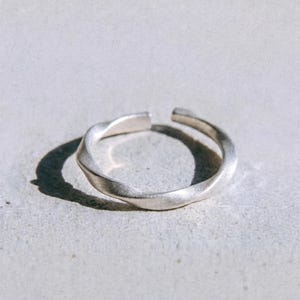 Puede incluir: Un anillo abierto de tono plateado con un diseño de banda trenzada y texturizada. El metal tiene un acabado mate y una abertura en la parte superior. La palabra dusk. está impresa en letras minúsculas negras en la parte inferior.