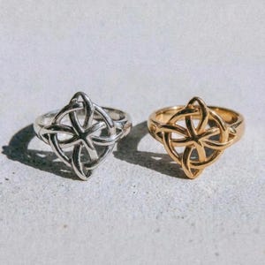 Op de afbeelding: Twee ringen met een Keltisch knoopontwerp. Eén ring is zilverkleurig, de andere goudkleurig. Ze worden gepresenteerd op een lichtgrijze ondergrond. Het woord "dusk." staat in het zwart onderaan de afbeelding.