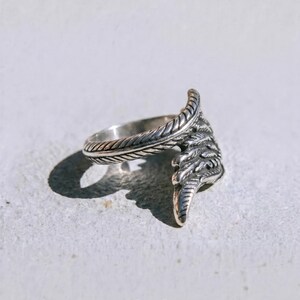 May include: A silver ring with a feather design. The ring has a detailed feather motif that wraps around the finger. The word "dusk." is printed at the bottom of the image.