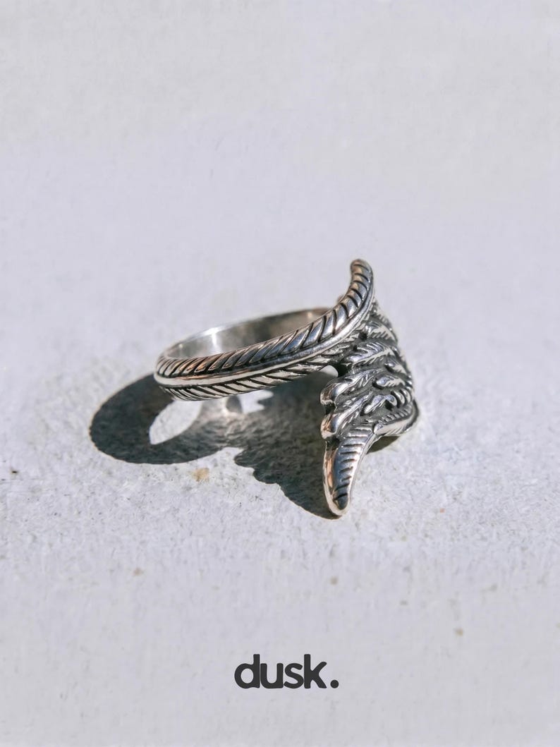 May include: A silver ring with a feather design. The ring has a detailed feather motif that wraps around the finger. The word "dusk." is printed at the bottom of the image.