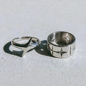 Puede incluir: Dos anillos de plata sobre una superficie gris claro. Uno tiene un diseño de estrella, y el otro es una banda gruesa con cortes de estrella. La palabra "dusk." está impresa en negro en la parte inferior.