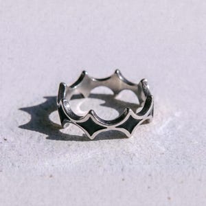 May include: A silver-toned metal ring featuring a repeating starburst or crown-like silhouette with black enamel inlay within each point. The band has a polished finish and the word "dusk." is printed in black text below the ring.