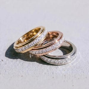May include: Three rings in gold, rose gold, and silver, each with a band of clear, square-cut stones. The rings are stacked, with the gold ring on top. The word "dusk." is printed in black at the bottom.