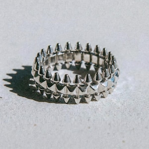 May include: A silver ring with a double row of conical spikes. The ring is made of a shiny metal and is set against a neutral background. The word "dusk." is printed at the bottom.
