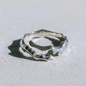 May include: A silver ring with an abstract, flowing design. The ring has an irregular shape with textured details and a polished finish. The word "dusk." is printed in a dark font at the bottom of the image.