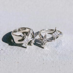 May include: Two silver rings with starburst designs, each featuring a central square of small, clear stones. The rings are displayed on a light grey surface. The word "dusk." is printed in black at the bottom.