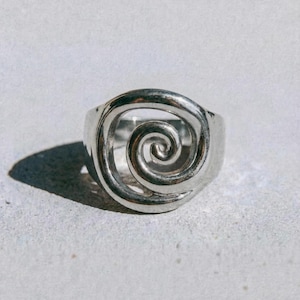 May include: A silver ring with a spiral design in the center. The ring has a thick band and a polished finish. The word "dusk." is printed in black at the bottom of the image.
