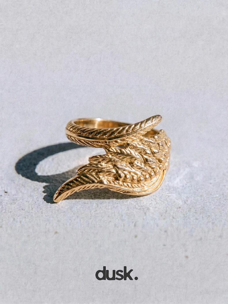 May include: A gold ring with a feather design. The ring has detailed texturing, resembling feathers. The word "dusk." is printed in black at the bottom of the image. The ring is a piece of jewellery.