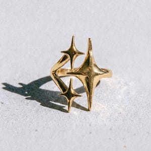 Può includere: Un anello dorato con un gruppo di disegni a forma di stella. L'anello ha un'estetica moderna e geometrica. La parola "dusk." è stampata in nero nella parte inferiore dell'immagine.
